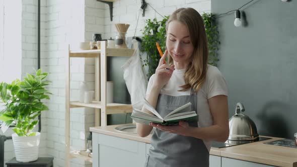 Young Woman Reading Cookbook in the Kitchen Looking for Recipe