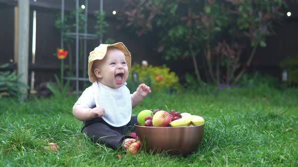 Small Newborn Child in Summer Panama Hat Sit on Grass Barefoot in Bib with Big Bowl of Fresh Fruit