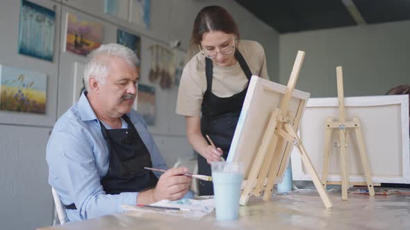 A Female Teacher Shows a Retired Man How to Draw a Picture with Paints and a Brush at Courses for