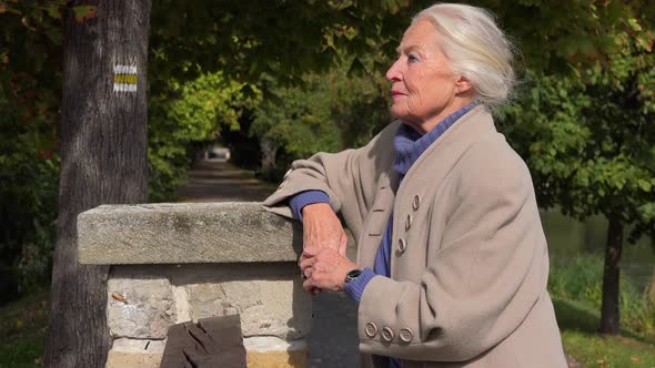 An Elderly Woman Stands on a Bridge, Leans Against a Stone Barrier and Looks Sad