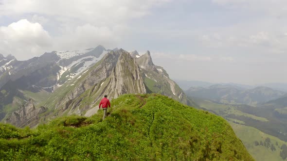 A man in a red jacket is walking on top of a mountain and is followed by the drone. There is a beaut