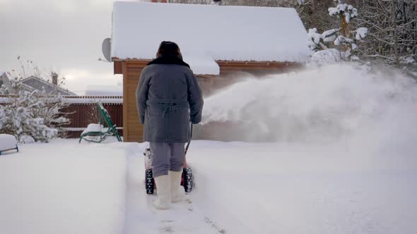 Man Cleans Snow With Snow Plow Background Of Wooden House In Winter