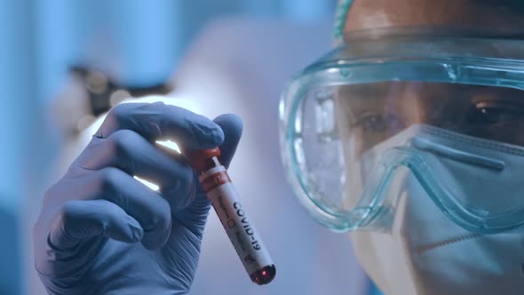 Doctor in Hazmat Suit and Protective Mask Holds a Test Tube with COVID-19 Virus