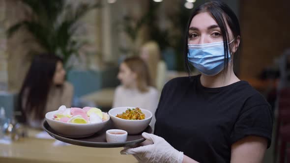 Confident Female Waiter in Coronavirus Face Mask Posing with Seafood on Tray and Blurred Clients