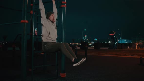 Bearded Guy Is Hanging on Training Area in Park in Night and Lifting Legs