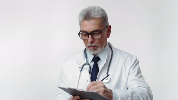 Elderly Bearded Therapist with Glasses Writes with a Pen on Paper and Then Looks at the Camera