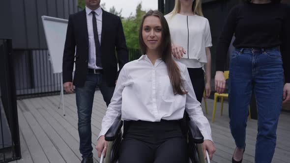 Portrait of Confident Disabled Woman in Wheelchair Smiling As Coworkers Approaching Putting Hands on