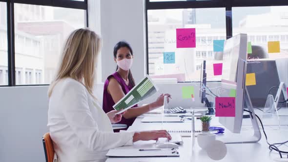 Diverse businesswomen wearing face masks using computers passing paperwork in office