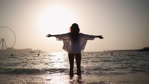 Woman Stands in Sea and Raises Her Arms to Sides on Background Sunset in Dubai