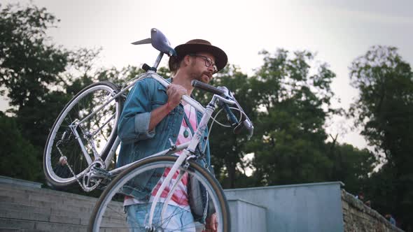 Young Stylish Hipster Man in Hat and Glasses Walking with His Bike in City Centre Holding It on