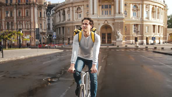 Portrait of Delivery Man with Yellow Backpack Riding Bike Through Old City Center with Beautiful Old