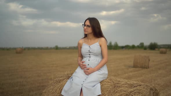 Young Pregnant Woman Sits on a Haystack in a Field