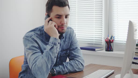 Young Office Worker Dialing a Number and Talking on His Mobile Phone at the Office Sitting at the