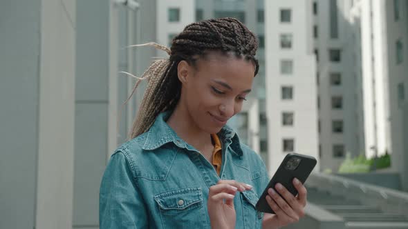 African American Woman with Dreadlocks and in a Denim Jacket Using Smart Phone Outdoors of the