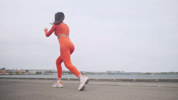 A woman in a bright orange fitness suit is doing exercises on the street. Pumps up the legs. A young
