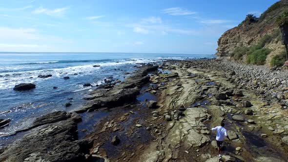 Tracking shot of a young man running on a rocky ocean beach shoreline.