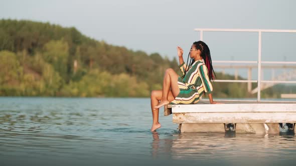 Beautiful AfricanAmerican Girl with Dreadlocks in a Bright Dress Sits on a Pier Among the Boats with