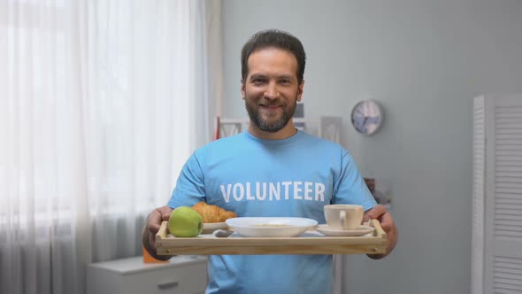 Middle-Aged Smiling Male Volunteer Showing Tray With Breakfast to Camera, Care