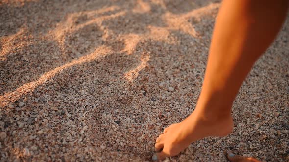 Female Leg of a Young Woman Draws a Heart on Sand and Pebbles on Beach