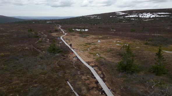 A woman and her dog walking on a mountain plank path with snow behind them. Fast aerial orbiting inf