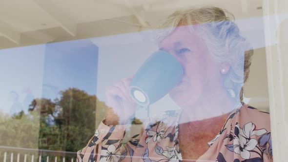 Caucasian senior woman drinking coffee while looking through the window at home