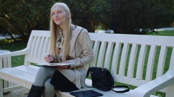 Beautiful Woman Sits on the Bench in Autumn Park, Makes Notes in a Notebook