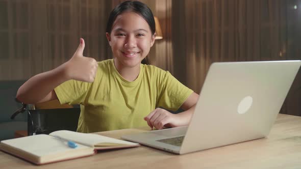 Asian Kid Girl Sitting In A Wheelchair Using Laptop Computer While Smiling And Thumb Up To Camera