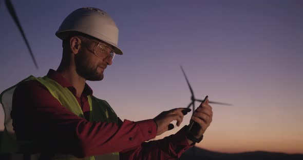 Engineer Browses Information in the Phone Standing on the Background of Windmills at Sunset