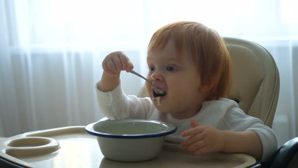 Close-up of a Baby Sitting at the Table and Eating Baby Food with a Spoon on Its Own