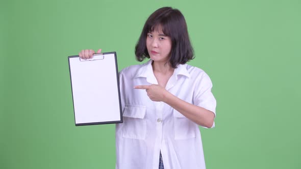 Stressed Asian Businesswoman Showing Clipboard and Giving Thumbs Down