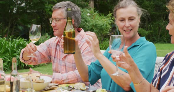 Animation of diverse happy senior female and male friends eating lunch in garden, pouring wine