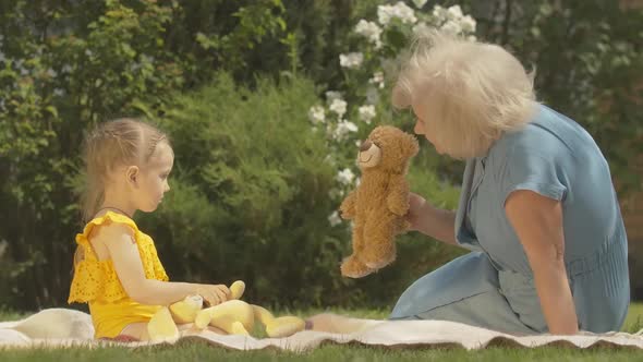 Side View of Positive Caucasian Senior Woman Playing Teddy Bear with Little Girl and Telling