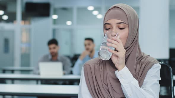 Thirsty Millennial Indian Arabic Female Worker in Hijab Holding Glass Drinking Pure Mineral Water in