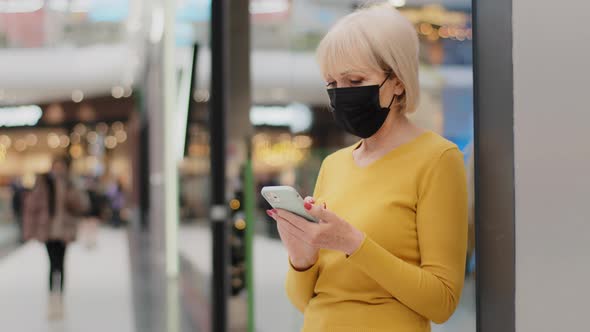 Mature Caucasian Woman in Protective Mask Stands in Shopping Center Holds Phone Communicates in