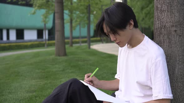 A Young Asian Male Student Sits in a Park and Writes in a Notebook