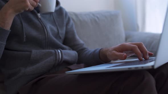 Young man drinks from a mug while typing on laptop at home