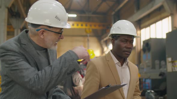 Multiethnic Engineers in Hardhats Walking and Speaking in Machinery Plant