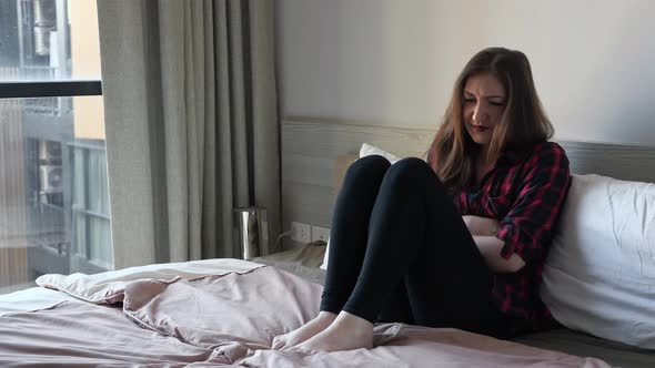 Young Woman Sits on Large Bed Writhes Holding Hands on Stomach