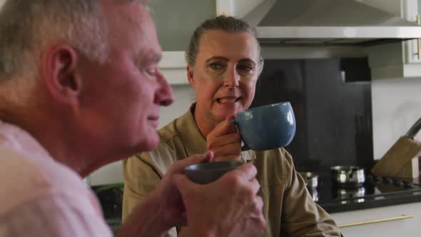 Caucasian senior couple holding coffee cup talking to each other in the kitchen at home