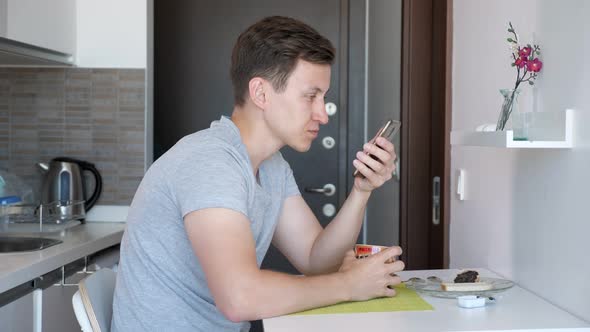 Man Is Having a Cup of Tea and Browsing Phone Sitting in a Small Kitchen