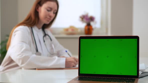 Laptop with Web Page Chromakey Mockup and Blurred Female Doctor Filling in Medical History at