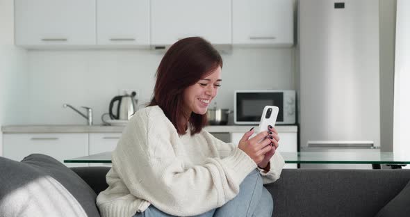 Young Woman Uses the Phone While Sitting on the Sofa in Modern Home Interior