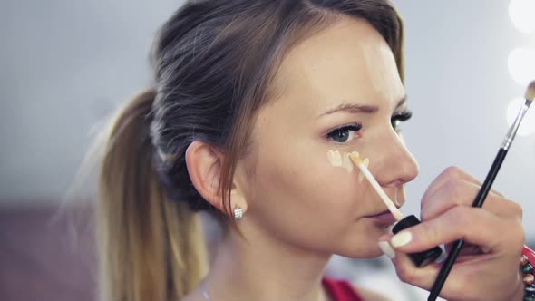 Closeup of Professional Makeup Artist Applying Concealer to Skin of Young Woman with Brush to Cover