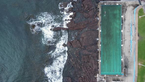 People exercising in a seaside swimming pool with waves crashing over coastal rock formation. High d