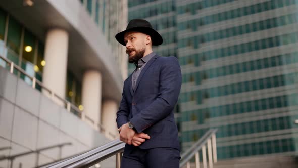 a Mustachioed and Bearded Stylish Man in a Plaid Suit and Hat Crossed Arms and Leans on the Railing