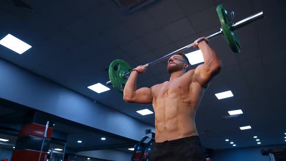 Handsome male bodybuilder lifts barbel over head. Selective focus on shirtless athlete. Slow motion.