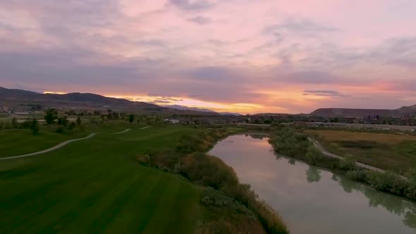 Aerial view of a golf course at sunset with a river below and the colorful sky reflecting off the ri