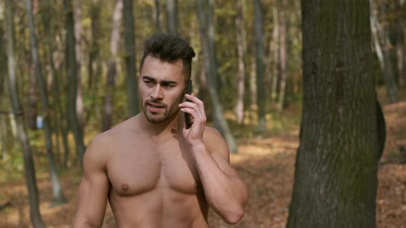 Strong Guy With Naked Torso Stands In Autumn Park And Speaks On Mobile Phone.