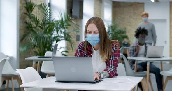 Portrait of Concentrated Young Caucasian Woman in Covid-19 Face Mask Working Online at Laptop in