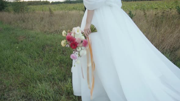 The Groom Across the Field Leads By the Hand the Bride in a Chic Dress with a Train and a Bouquet of